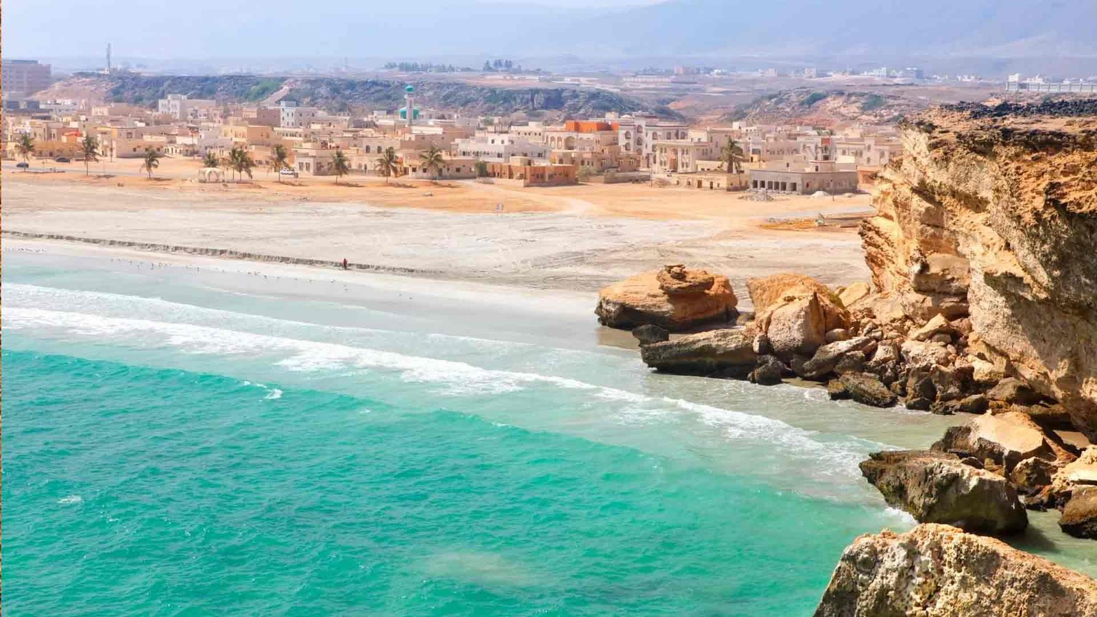 Beautiful coastal view of a sandy beach with turquoise waters and a backdrop of traditional Omani buildings, representing one of the serene Oman tourist attractions