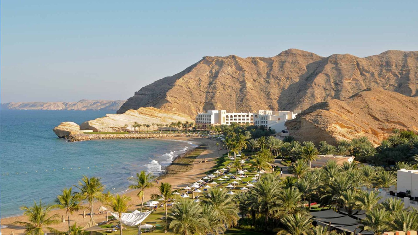 stunning coastal view of Oman with a pristine beach, lush palm trees, and a luxurious resort nestled against rugged mountains, illustrating the ideal weather during the best time to visit Oman.