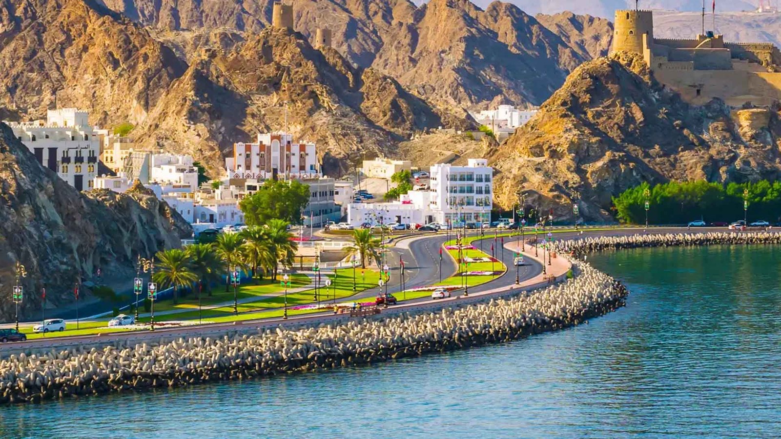 A scenic view of Muscat, Oman, with traditional architecture nestled between rugged mountains and a coastal road, highlighting the blend of natural beauty and cultural heritage integral to Eco-Tourism in Oman