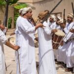 A group of Omani men dressed in traditional white dishdashas participate in a cultural dance, holding swords and drums. The scene is vibrant, with the men engaged in singing and drumming, reflecting the rich cultural heritage of Oman. This image captures the essence of "Oman cultural etiquette and traditions."