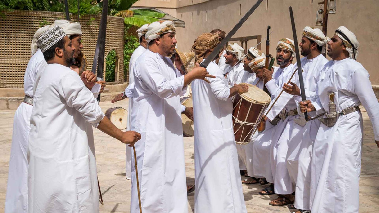 A group of Omani men dressed in traditional white dishdashas participate in a cultural dance, holding swords and drums. The scene is vibrant, with the men engaged in singing and drumming, reflecting the rich cultural heritage of Oman. This image captures the essence of "Oman cultural etiquette and traditions."