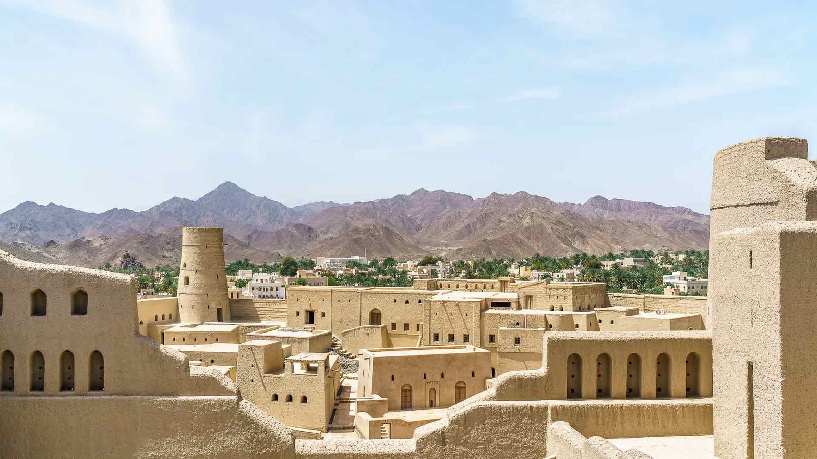 Panoramic view of Nizwa Fort in Oman, with its historic mud-brick walls and watchtowers set against a backdrop of rugged mountains and a clear blue sky. The image captures the fort's significance as a cultural landmark, highlighting the architectural beauty and historical depth of the region. The best time to visit Oman and explore such inland gems is during the cooler winter months, when the weather is ideal for outdoor exploration.