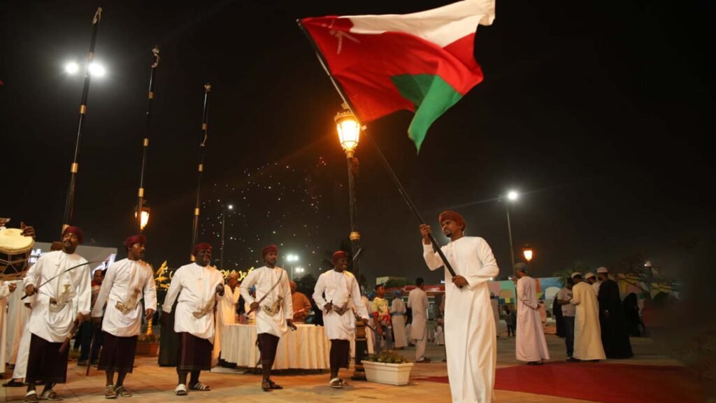 A group of Omani men in traditional attire performing a cultural dance at a night event, with one man proudly waving the Omani flag, showcasing the rich traditions highlighted in the "Omani Festivals and Events"