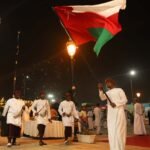 A group of Omani men in traditional attire performing a cultural dance at a night event, with one man proudly waving the Omani flag, showcasing the rich traditions highlighted in the "Omani Festivals and Events"