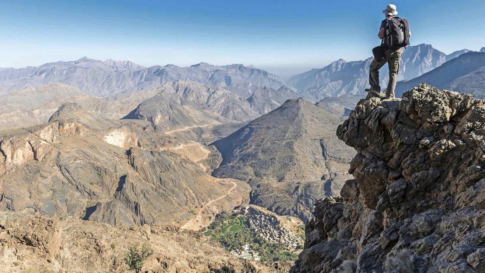 A hiker stands on a rocky outcrop overlooking a vast, rugged mountain landscape in the Al Hajar range. The expansive view reveals winding trails, steep cliffs, and a distant village nestled in the valley. This image epitomizes the adventurous spirit and breathtaking scenery found along Oman Hiking Trails, offering an unforgettable experience for those exploring the region’s natural beauty.