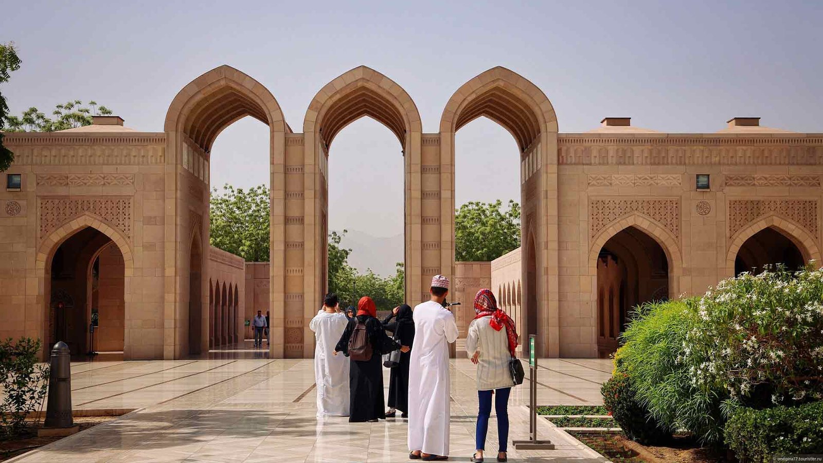 Visitors exploring the entrance of the Sultan Qaboos Grand Mosque in Muscat, Oman, with its intricate Islamic architecture and beautifully landscaped surroundings. The arches and pathways reflect the elegance and cultural significance of the site. The best time to visit Oman, particularly to enjoy outdoor sites like this mosque, is during the cooler winter months, when the weather is mild and pleasant for sightseeing.