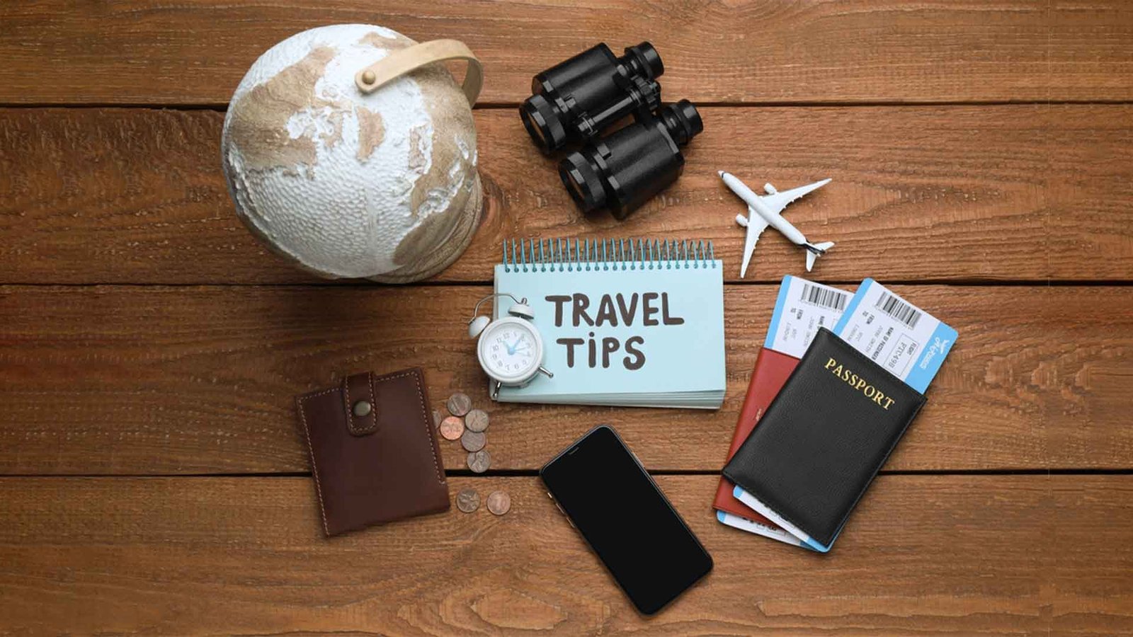 A flat lay image featuring travel essentials on a wooden surface, including a globe, binoculars, a small airplane model, a passport with travel tickets, a notebook labeled "Travel Tips," an alarm clock, a smartphone, coins, and a leather wallet. The image reflects the importance of preparation and safety for travelers, particularly those seeking "Oman travel safety tips" for a secure and well-planned journey.