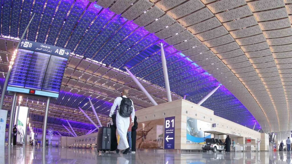 A traveler walks through a modern, spacious terminal at an Omani airport, surrounded by purple-lit ceilings and clear signage pointing to check-in zones. The scene highlights the well-organized and welcoming atmosphere for those navigating Oman airport transportation guide.